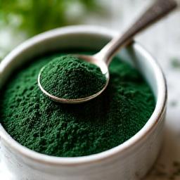 Serbuk spirulina hijau tua di dalam bekas kecil dengan sudu, siap ditambah ke minuman.